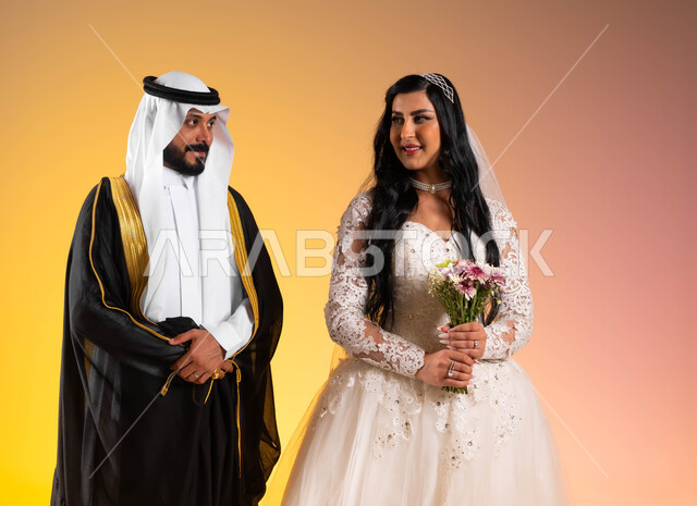 أصالة الأعراس في المملكة، مفهوم التزيين والاطلالة المتأنقة، بورتريه مقرب لعروس عربية خليجية سعودية مبتسمة ترتدي فستان الزفاف الأبيض تحمل بيدها باقة ورد، عريس ينظر الى زوجته بتعابير المودة, نظرات مليئة بالرومنسية والحب، خلفية ملونة