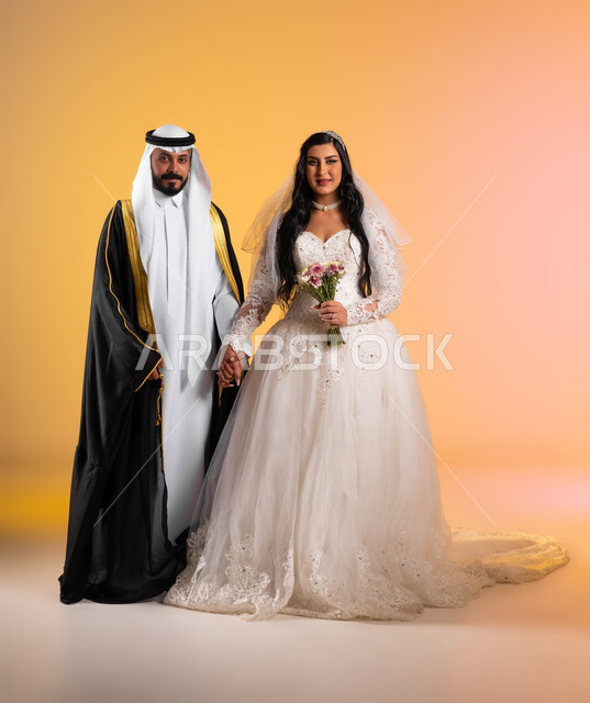 النظر الى الكاميرا بتعابير السرور والفرح بليلة العمر، مفهوم الاستقرار وتكوين عائلة، بورتريه لعريس عربي خليجي سعودي مبتسم يرتدي البشت والغترة التقليدية يقف بجانب زوجته، عروس ترتدي فستان الزفاف الأبيض تحمل بيدها باقة ورود ملونة، الجسم بالطول الكامل، خلفية ملونة