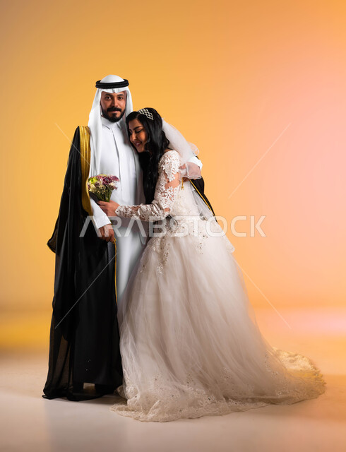 عناق مليء بالرومنسية والحب والأمان، مراسم الأعراس السعودية، مفهوم التزيين والاطلالة المتألقة، بورتريه لعروس عربية خليجية سعودية مبتسمة ترتدي فستان الزفاف الأبيض تحمل بيدها باقة ورد تقوم بمعانقة عريسها بتعابير المودة, صورة لكامل الجسم، خلفية ملونة