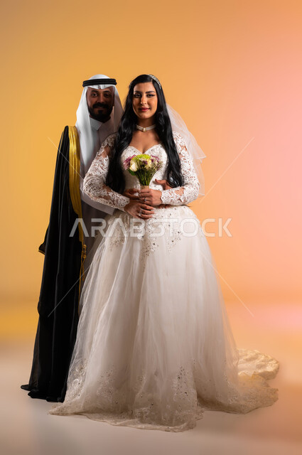 عناق مليء بالرومنسية والحب، مراسم الأعراس في المملكة، مفهوم التزيين والاطلالة المتألقة والمظهر الخارجي، بورتريه لعروس عربية خليجية سعودية مبتسمة ترتدي فستان الزفاف الأبيض تحمل بيدها باقة ورد تقف أمام عريسها بتعابير المودة, صورة لكامل الجسم، خلفية ملونة