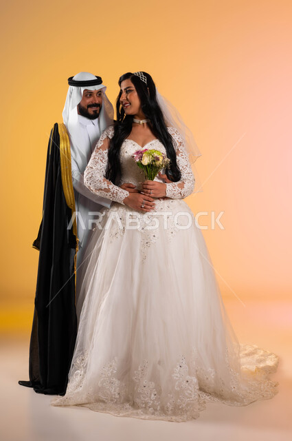 تبادل نظرات مليئة بالرومنسية والحب، فخامة الأعراس في المملكة، مفهوم التزيين والاطلالة المتأنقة، بورتريه لعروس عربية خليجية سعودية مبتسمة ترتدي فستان الزفاف الأبيض تحمل بيدها باقة ورد، عريس ينظر الى زوجته بتعابير المودة, الطول الكامل للجسم، خلفية ملونة