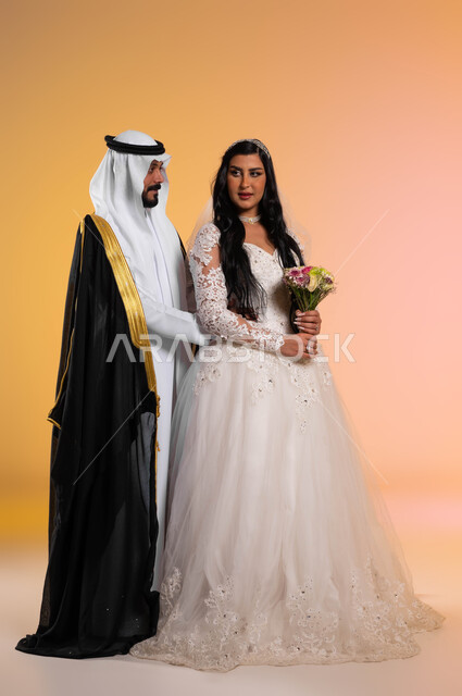 نظرات مليئة بالرومنسية والحب، فخامة الأعراس في المملكة، مفهوم التزيين والاطلالة المتأنقة، بورتريه لعروس عربية خليجية سعودية مبتسمة ترتدي فستان الزفاف الأبيض تحمل بيدها باقة ورد، عريس ينظر الى زوجته بتعابير المودة, الطول الكامل للجسم، خلفية ملونة