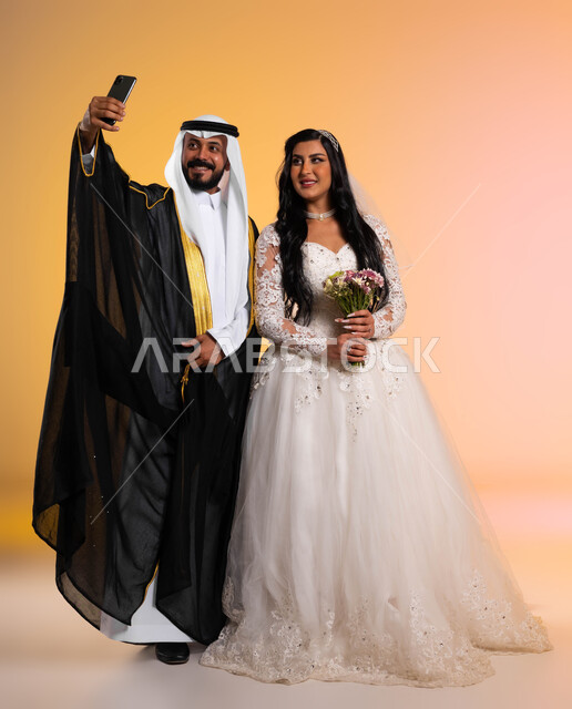 الاحتفال بإيماءات البشاشة والسرور في يوم الزفاف، استخدام الأجهزة والتقنيات الحديثة، بورتريه لعريس عربي خليجي سعودي يرتدي البشت والغترة التقليدية يقوم بالتقاط صور سيلفي تذكارية عبر الهاتف المحمول برفقة زوجته، الأناقة والاهتمام بالمظهر الخارجي، الطول الكامل للجسم، خلفية ملونة