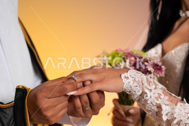 مراسم الاعراس والمناسبات في المملكة، استبدال الخواتم في ليلة الزفاف، بورتريه مقرب لرجل عربي خليجي سعودي يرتدي البشت والغترة التقليدية يقوم بتلبيس الخاتم لعروسته، عروس سعودية ترتدي الفستان الأبيض وتحمل بيدها باقة الورود، خلفية ملونة