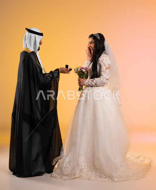 الأعراس والمناسبات الراقية في المملكة, الاستعداد للاحتفال في ليلة العمر، بورتريه من الجانب لعريس عربي خليجي سعودي يرتدي البشت والغترة التقليدية يقوم بإهداء خاتم الزفاف لزوجته، عروس مبتسمة تقف بجانب زوجها بايماءات المفاجئة والسرور، الطول الكامل للجسم، خلفية ملونة