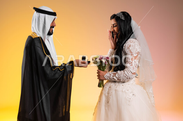 الاستعداد للاحتفال في ليلة العمر، المناسبات والأعراس الراقية في المملكة, بورتريه من الجانب لعريس عربي خليجي سعودي يرتدي البشت والغترة التقليدية يقوم بإهداء خاتم الزفاف لزوجته، عروس مبتسمة تقف بجانب زوجها بايماءات المفاجئة والسرور، خلفية ملونة