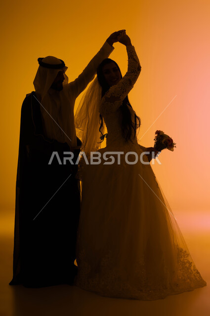 الحب والمودة بين الزوجين، مفهوم الاستقرار وتكوين الأسرة، بورتريه ظلي لعريس يقوم بالرقص مع عروسته بايماءات البشاشة في يوم الزواج، عروس عربية خليجية سعودية  ترتدي فستان الزفاف الأبيض والطرحة تحمل بيدها باقة الورد، صورة الجسم بالطول الكامل، خلفية ملونة، سالويت