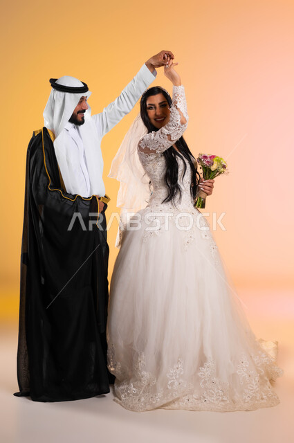 مفهوم الاستقرار وتكوين الأسرة، الحب والمودة بين الزوجين، بورتريه لعريس يقوم بالرقص مع عروسته بايماءات البشاشة في يوم الزواج، عروس عربية خليجية سعودية  ترتدي فستان الزفاف الأبيض والطرحة تحمل بيدها باقة الورد، صورة الجسم بالطول الكامل، خلفية ملونة