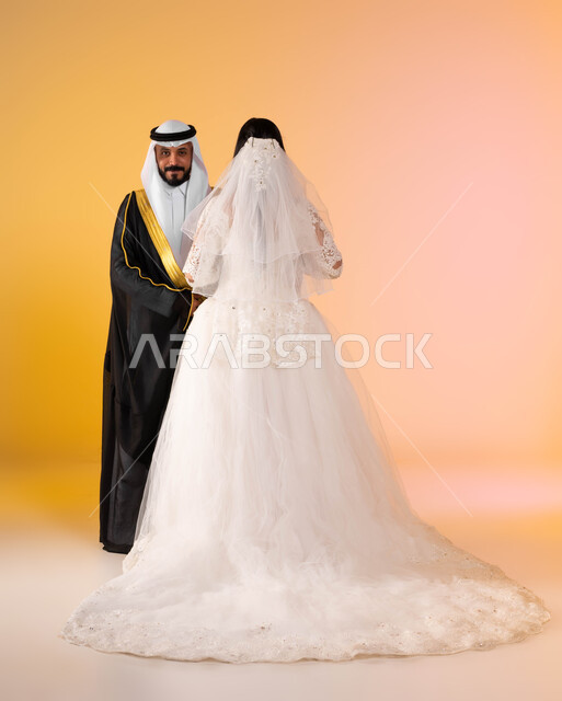 الاستعداد للاحتفال بليلة العمر، مراسم الاعراس في المملكة، بورتريه لعريس عربي خليجي سعودي مبتسم ينظر الى الكاميرا، عروس سعودية ترتدي فستان الزفاف والطرحة البيضاء تقف جانب زوجها في حفل العرس، الاهتمام بالمظهر الخارجي، الطول الكامل للجسم، خلفية ملونة