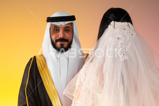 مراسم الاعراس والزواج في المملكة، الاستعداد للاحتفال بليلة العمر، بورتريه مقرب لعريس عربي خليجي سعودي مبتسم ينظر الى الكاميرا، عروس سعودية ترتدي فستان الزفاف والطرحة البيضاء تقف بجانب زوجها في حفل العرس، الاهتمام بالمظهر الخارجي، خلفية ملونة