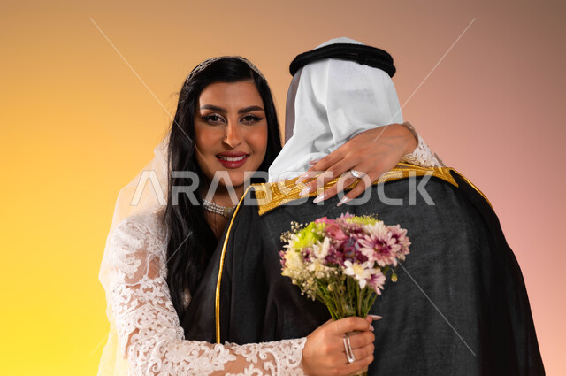 تكوين أسرة وتحقيق الاستقرار العاطفي، ايماءات الفرح والسرور في يوم الزواج، بورتريه مقرب لعروس عربية خليجية سعودية مبتسمة ترتدي فستان الزفاف الأبيض تحمل بيدها باقة ورد وتقوم بمعانقة زوجها بحب, لحظات مليئة بالمحبة والود في حفل الزفاف، خلفية ملونة