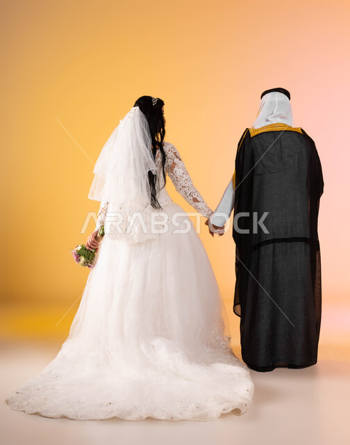 الاهتمام بالاناقة والمظهر الخارجي، الاستعداد للاحتفال في ليلة العمر، بورتريه من الخلف لعريس وعروس عربيين خليجين سعوديين يستمتعان باللحظات والأجواء السعيدة في يوم الزواج، ارتداء أزياء الأعراس التقليدية في يوم الزفاف، الجسم بالطول الكامل، خلفية ملونة