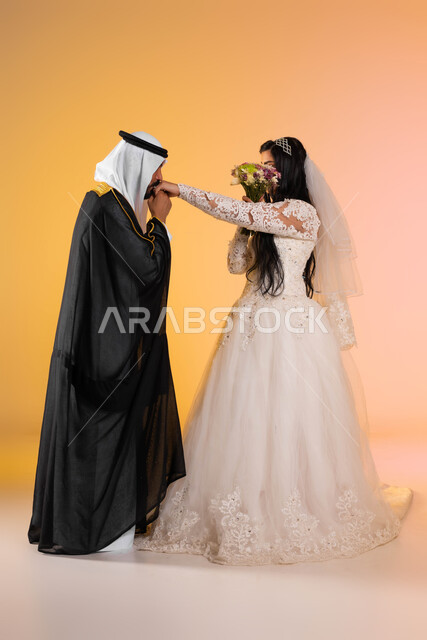 مفهوم الخجل والحياء عند النساء، بورتريه لزوجان عربيان خليجيان في ليلة الزفاف, عروس تغطي وجهها بباقة الورد حياء وخجلا، عريس يرتدي البشت والغترة التقليدية يقبل يد عروسته بحب واحترام، الطول الكامل للجسم، خلفية ملونة