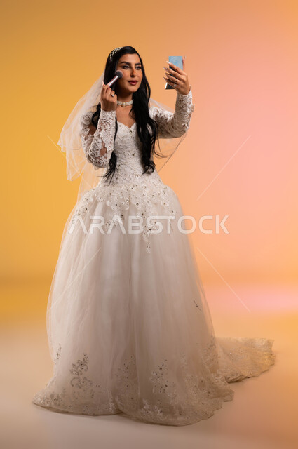 ايماءات النظر إلى المرآة بتعابير البهجة والسعادة، استعمال مساحيق التجميل لإبراز الجمال، مفهوم التزيين والاطلالة الأنيقة، بورتريه لعروس عربية سعودية خليجية ترتدي فستان الزفاف الأبيض تحمل بيدها المرآة وتقوم بوضع احمر الخدود، صورة الجسم بالطول الكامل، خلفية ملونة