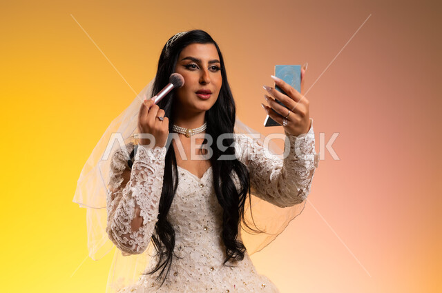 النظر إلى المرآة بتعابير البهجة والسعادة، استعمال مساحيق التجميل لإبراز الجمال، مفهوم التزيين والاطلالة الأنيقة، بورتريه لعروس عربية سعودية خليجية ترتدي فستان الزفاف الأبيض تحمل بيدها المرآة وتقوم بوضع احمر الخدود، خلفية ملونة