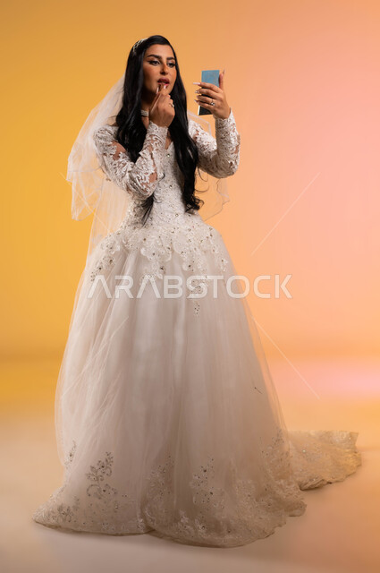 الاستعداد للاحتفال بليلة العمر،مفهوم التزيين والإطلالة المتألقة، بورتريه لعروس عربية خليجية سعودية ترتدي فستان الزفاف الأبيض وتنظر لنفسها بالمرآة وتقوم بوضع أحمر الشفاه، استخدام مساحيق التجميل لإبراز الجمال، صورة الجسم بالطول الكامل، خلفية ملونة