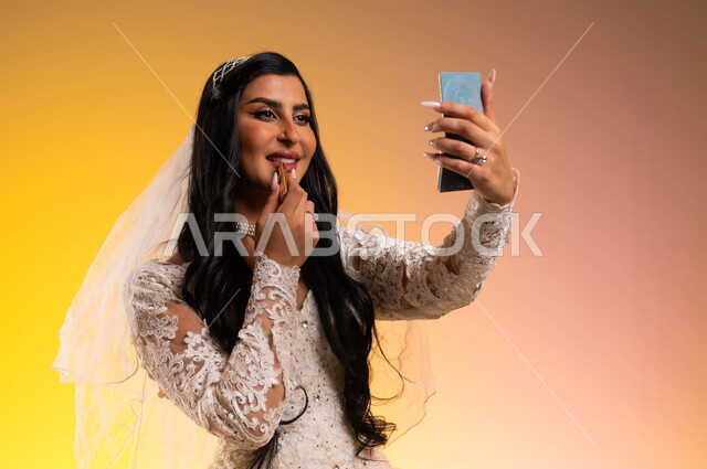 الاهتمام بالمظهر الخارجي، مفهوم التزيين والإطلالة المتألقة، بورتريه مقرب لعروس عربية خليجية سعودية ترتدي فستان الزفاف الأبيض تنظر لنفسها لالمرآة وتقوم بوضع أحمر الشفاه، استخدام مساحيق التجميل لإبراز الجمال، الاستعداد للاحتفال بليلة العمر، خلفية ملونة