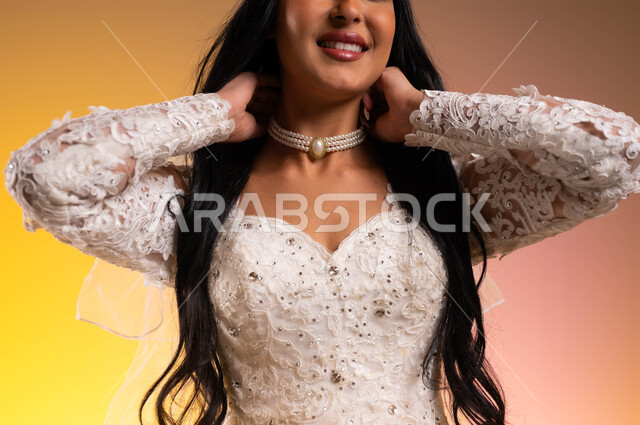 الاهتمام بالأناقة والمظهر الخارجي، مراسم الأعراس والزواج في السعودية، بورتريه مقرب لعروسة عربية خليجية سعودية ترتدي بدلة الزفاف البيضاء تقوم بارتداء عقد مرصع بالألماس، ايماءات السعادة والسرور، مفهوم التزين والاطلالة المتألقة، خلفية ملونة