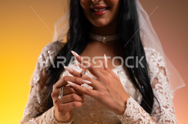 الاهتمام بالأناقة والمظهر الخارجي، بورتريه مقرب لعروس عربية خليجية سعودية ترتدي فستان الزفاف الأبيض تقوم بارتداء خاتم زواج من ألماس، الاستعداد من أجل ليلة العمر، ايماءات السعادة والسرور، مفهوم التأنق والتجمل، خلفية ملونة