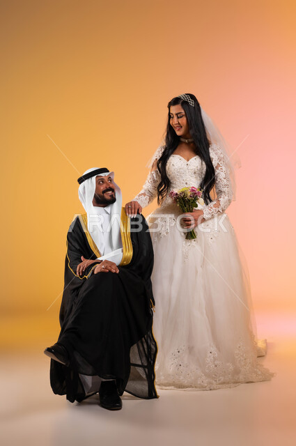 ايماءات الفرح والسرور يوم الزفاف، تبادل نظرات الحب والمودة، بورتريه لعريس عربي خليجي سعودي يجلس ويرتدي البشت والغترة التقليدية، عروس سعودية ترتدي فستان الزفاف الأبيض تضع يدها على كتف زوجها، مراسم الأعراس والزواج في السعودية، الجسم بالطول الكامل، خلفية ملونة