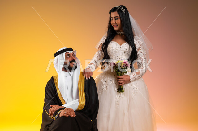 تبادل نظرات الحب والمودة، بورتريه لعريس عربي خليجي سعودي يجلس ويرتدي البشت والغترة التقليدية، عروس سعودية ترتدي فستان الزفاف الأبيض تضع يدها على كتف زوجها، ايماءات الفرح والسرور يوم الزفاف، مراسم الأعراس والزواج في السعودية، خلفية ملونة