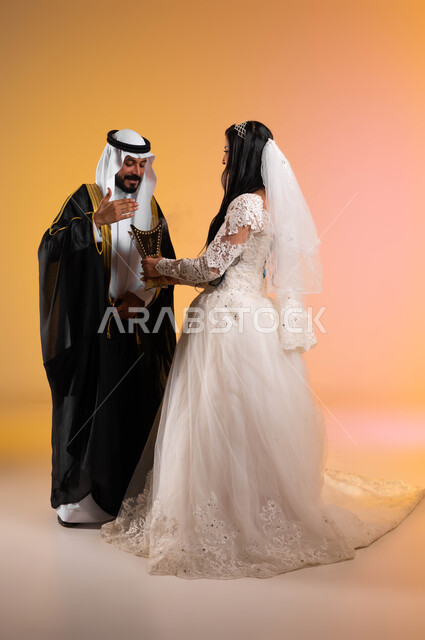 التطيب بالمسك والعود، مفهوم الاستقرار وتكوين عائلة، بورتريه من الجانب لعروس عربية خليجية سعودية تحمل بيدها المبخرة وتعطر زوجها، ارتداء الأزياء الخليجية الفاخرة، مراسم الأعراس والمناسبات في المملكة، الاهتمام بالمظهر الخارجي، الطول الكامل للجسم، خلفية ملونة