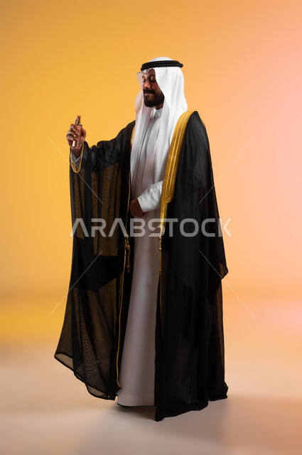 الأناقة والاهتمام بالمظهر الخارجي، بورتريه لرجل عربي خليجي سعودي يرتدي البشت والغترة التقليدية يقوم برش العطر، استخدام الزيوت العطرية الطبيعية، عطور ذات روائح فاخرة، مراسم الأعراس والزواج في السعودية، صورة الجسم بالطول الكامل، خلفية ملونة