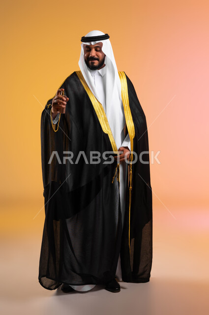 مفهوم الأناقة والاهتمام بالمظهر الخارجي، بورتريه لرجل عربي خليجي سعودي يرتدي البشت والغترة التقليدية يقوم برش العطر، استخدام الزيوت العطرية الطبيعية، عطور ذات روائح فاخرة، مراسم الأعراس والزواج في السعودية، صورة الجسم بالطول الكامل، خلفية ملونة
