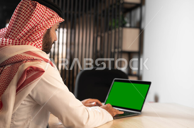 دور التكنولوجيا في في تسهيل الأعمال، استعمال الحاسوب المحمول في إنجاز العمل، تصفح الانترنت باستخدام الأجهزة الحديثة، صورة مقربة من الجانب لرجل عربي خليجي سعودي يرتدي الزي التقليدي والشماغ يقوم بالنقر على جهاز اللابتوب بشاشة خضراء فارغة ، كروما