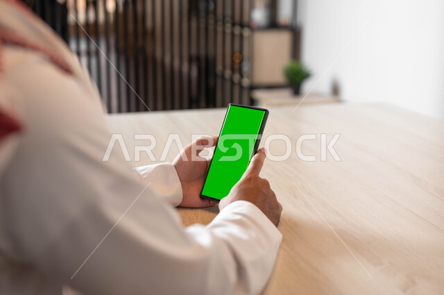 ادارة الأعمال وعقد الاجتماعات، تصفح الانترنت باستخدام الجوال، دمج التكنولوجيا الحديثة بالحياة العملية، انجاز مهام العمل عن بعد، صورة مقربة من الخلف لرجل عربي خليجي سعودي يرتدي الزي التقليدي يحمل بيده الهاتف المحمول بشاشة خضراء فارغة، كروما