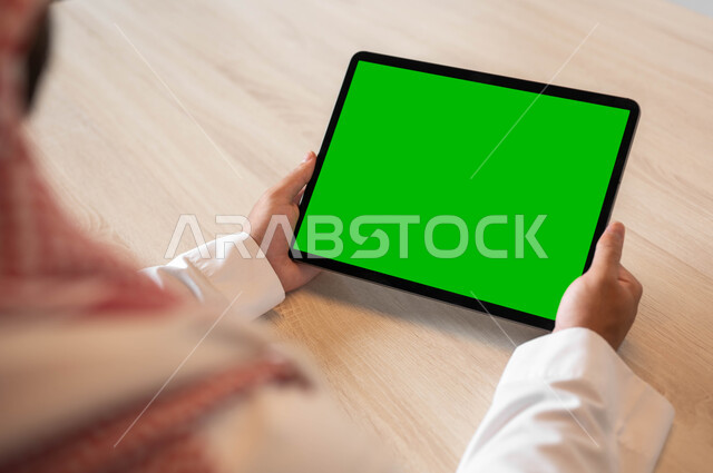 تصفح مواقع التواصل الاجتماعي عن طريق الانترنت، استخدام الأجهزة الحديثة في انجاز مهام الأعمال، استعمال التكنولوجيا في العمل عن بعد، صورة مقربة من الخلف لرجل أعمال عربي خليجي سعودي يرتدي الزي التقليدي والشماغ يحمل بيده حهاز التابليت بشاشة خضراء فارغة، كروما