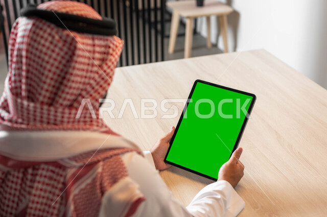 استعمال التكنولوجيا في العمل عن بعد، تصفح مواقع التواصل الاجتماعي عن طريق الانترنت، استخدام الأجهزة الحديثة في انجاز مهام الأعمال، صورة مقربة من الخلف لرجل عربي خليجي سعودي يرتدي الزي التقليدي والشماغ يحمل بيده جهاز التابلت بشاشة خضراء فارغة، ، كروما