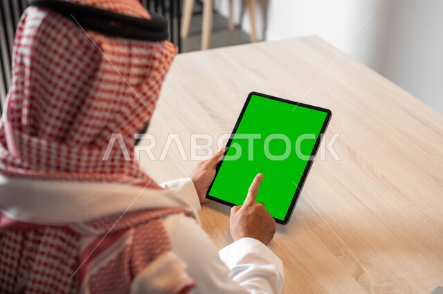 Holding meetings via the Internet, completing work quickly and easily, using modern technology in remote work, technical development in various fields, a close-up photo from the back of a Saudi Gulf Arab man wearing traditional clothing and a shemagh, holding a tablet in his hand with a blank green screen, chroma