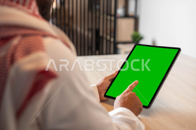 استخدام الجهاز اللوحي للعمل عن بعد، إنجاز الأعمال بسرعة وسهولة، عقد الاجتماعات عن طريق الانترنت، التطور التقني والتكنولوجي في مختلف المجالات، صورة مقربة من الخلف لرجل عربي خليجي سعودي يضع على رأسه الشماغ يحمل بيده التابليت، شاشة خضراء فارغة، كروما