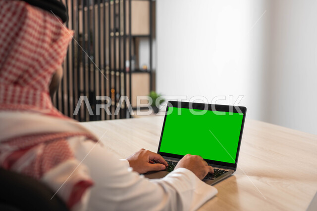 دور التكنولوجيا في في تسهيل الأعمال، التطور التقني والتكنولوجي في مختلف المجالات، تصفح الانترنت باستخدام الأجهزة الحديثة، صورة مقربة من الجانب لرجل عربي خليجي سعودي يرتدي الزي التقليدي والشماغ يقوم بالنقر على لابتوب بشاشة خضراء فارغة، كروما