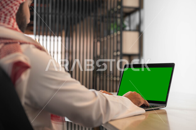 تصفح الانترنت باستخدام الأجهزة الحديثة، استعمال الحاسوب المحمول في إنجاز العمل، دور التكنولوجيا في في تسهيل الأعمال، تصورة مقربة من الجانب لرجل عربي خليجي سعودي يرتدي الزي التقليدي والشماغ يقوم بالنقر على لابتوب بشاشة خضراء فارغة، كروما