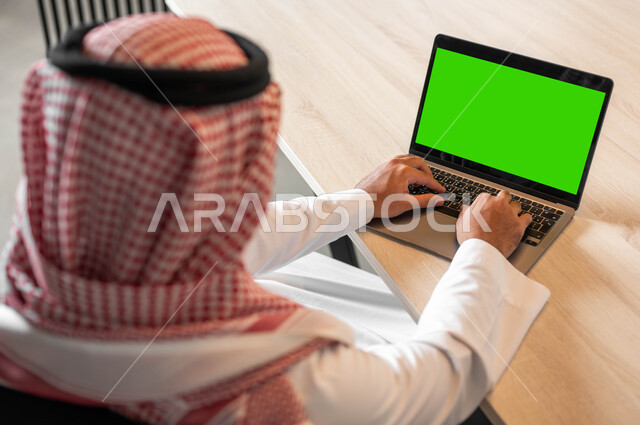 استعمال الحاسوب المحمول في إنجاز العمل، دور التكنولوجيا في في تسهيل الأعمال، تصفح الانترنت باستخدام الأجهزة الحديثة، صورة مقربة من الجانب لرجل عربي خليجي سعودي يضع على رأسه الشماغ يقوم بالنقر على لابتوب بشاشة خضراء فارغة، كروما