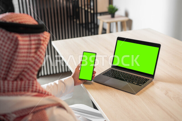 استعمال اللابتوب لادارة الأعمال وعقد الاجتماعات عن بعد، تصفح الانترنت باستخدام الجوال، دمج التكنولوجيا الحديثة بالحياة العملية، صورة مقربة من الخلف لرجل عربي خليجي سعودي يرتدي الزي التقليدي والشماغ يحمل بيده الهاتف المحمول بشاشة خضراء فارغة، كروما