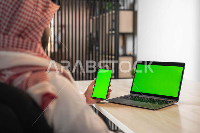 عقد الاجتماعات عن بعد، تصفح الانترنت باستخدام الجوال، دمج التكنولوجيا الحديثة بالحياة العملية، استعمال اللابتوب لادارة الأعمال، صورة مقربة من الخلف لرجل عربي خليجي سعودي يرتدي الزي التقليدي والشماغ يحمل بيده الهاتف المحمول بشاشة خضراء فارغة، كروما