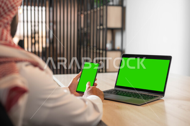 صفح الانترنت باستخدام الجوال، دمج التكنولوجيا الحديثة بالحياة العملية، استعمال اللابتوب لادارة الأعمال وعقد الاجتماعات عن بعد، صورة مقربة من الخلف لرجل عربي خليجي سعودي يرتدي الزي التقليدي والشماغ يحمل بيده الهاتف المحمول بشاشة خضراء فارغة، كروما