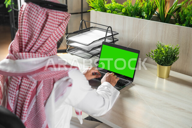 تصفح الانترنت باستخدام الأجهزة التقنية الحديثة، دمج التكنولوجيا الحديثة بالحياة العملية، إنجاز الأعمال عن بعد، صورة مقربة من الخلف لرجل عربي خليجي سعودي يرتدي الزي التقليدي والشماغ يقوم بالكتابة على لابتوب بشاشة خضراء فارغة، كروما