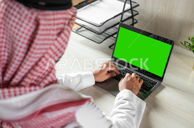 إنجاز الأعمال عن بعد، دمج التكنولوجيا الحديثة بالحياة العملية، تصفح الانترنت باستخدام الأجهزة التقنية الحديثة، صورة مقربة من الخلف لرجل عربي خليجي سعودي يرتدي الزي التقليدي والشماغ يقوم بالنقر على أزرار لابتوب بشاشة خضراء فارغة، كروما