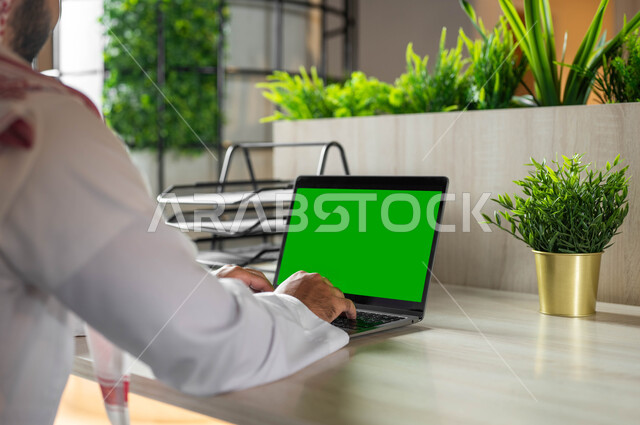 Integrating modern technology into practical life, doing work remotely, viewing the laptop screen, browsing the Internet using modern technical devices, a close-up photo from the back of the hands of a Saudi Gulf Arab man writing on a laptop with a blank green screen, chroma