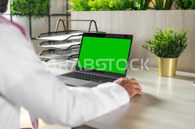 ادارة الأعمال و عقد الاجتماعات عن بعد، تصفح الانترنت باستخدام الأجهزة التقنية الحديثة، دمج التكنولوجيا الحديثة بالحياة العملية، صورة مقربة من الخلف لرجل عربي خليجي سعودي يرتدي الزي التقليدي يجلس على مكتب أمام لابتوب بشاشة خضراء فارغة، كروما