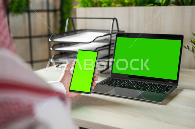 دمج التكنولوجيا الحديثة بالحياة العملية، استعمال اللابتوب لادارة الأعمال وعقد الاجتماعات عن بعد، تصفح الانترنت باستخدام الجوال، صورة مقربة من الخلف لرجل عربي خليجي سعودي يرتدي الزي التقليدي والشماغ يحمل بيده الهاتف المحمول بشاشة خضراء فارغة، كروما