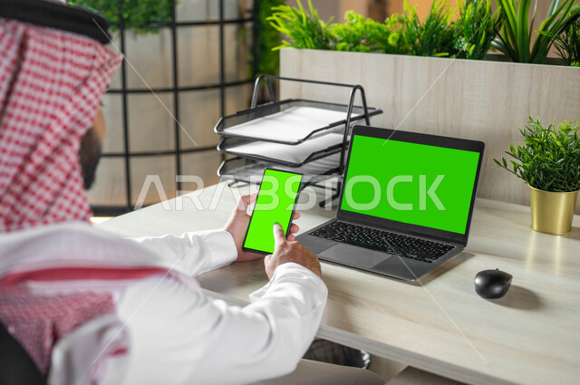 استخدام اللابتوب لادارة الأعمال وعقد الاجتماعات عن بعد، تصفح الانترنت باستعمال الجوال، دمج التكنولوجيا الحديثة بالحياة العملية، صورة مقربة من الخلف لرجل عربي خليجي سعودي يرتدي الثوب التقليدي والشماغ يحمل بيده الهاتف المحمول بشاشة خضراء فارغة، كروما