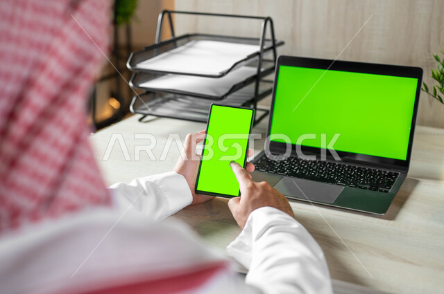 استعمال اللابتوب لادارة الأعمال وعقد الاجتماعات عن بعد، تصفح الانترنت باستخدام الجوال، دمج التكنولوجيا الحديثة بالحياة العملية، صورة مقربة من الخلف لرجل عربي خليجي سعودي يرتدي الزي التقليدي والشماغ يحمل بيده الهاتف المحمول بشاشة خضراء فارغة، كروما