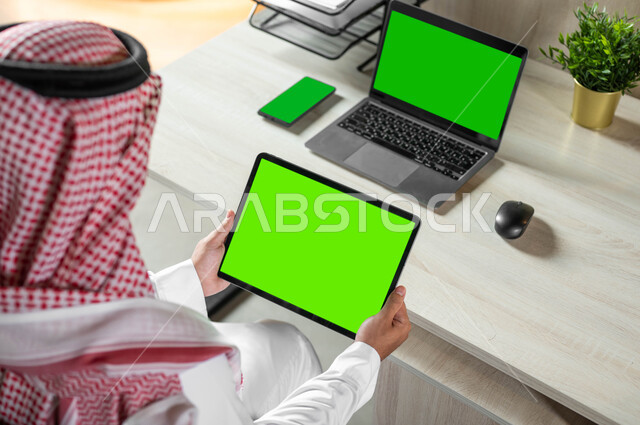Using electronic devices to accomplish daily tasks, using a laptop to organize work plans, technical and technological development in various fields, a close-up photo from the back of a Saudi Gulf Arab man holding a tablet with a blank green screen, chroma