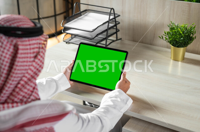 عقد الاجتماعات عن طريق الانترنت، إنجاز الأعمال بسرعة وسهولة، استخدام الجهاز اللوحي للعمل عن بعد، التطور التقني والتكنولوجي في مختلف المجالات، صورة مقربة من الخلف لرجل عربي خليجي سعودي يرتدي الزي التقليدي والشماغ يحمل بيده تابليت بشاشة خضراء فارغة، كروما