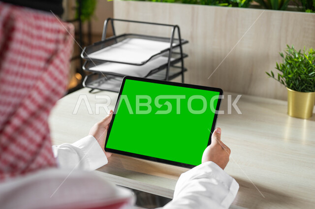 إنجاز الأعمال بسرعة وسهولة، استخدام الجهاز اللوحي للعمل عن بعد، عقد الاجتماعات عن طريق الانترنت، التطور التقني والتكنولوجي في مختلف المجالات، صورة مقربة من الخلف لرجل عربي خليجي سعودي يرتدي الزي التقليدي والشماغ يحمل بيده التابليت بشاشة خضراء فارغة، كروما